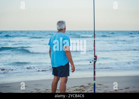 Un homme aîné dans une chemise bleue se tient contemplativement à côté de sa canne à pêche sur les rives sablonneuses de Valence, en Espagne, alors que les vagues s'écrasent contre la plage dure Banque D'Images