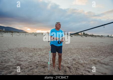 Un pêcheur plus âgé se tient sur une plage de sable à Valence, tenant une canne à pêche au coucher du soleil son regard concentré intentionnellement sur l'horizon, il attend une prise Banque D'Images