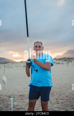 Un homme âgé se tient fièrement avec sa canne à pêche sur une plage de sable à Valence, en Espagne, lors d'un coucher de soleil serein ses vêtements de plage décontractés et attentionnés Banque D'Images