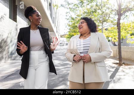 Deux femmes professionnelles appréciant une conversation tout en marchant dans une rue ensoleillée de la ville. La femme afro-américaine fait des gestes animés comme les deux rires toge Banque D'Images