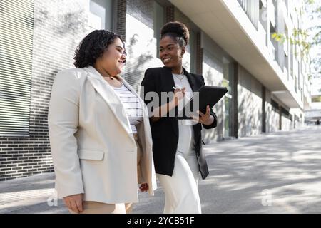 Deux femmes d’affaires afro-américaines s’engagent dans une discussion tout en marchant à l’extérieur, l’une tenant une tablette numérique. Les deux sont souriants et professionnellement dr. Banque D'Images