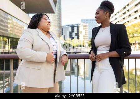 Une femme professionnelle afro-américaine et hispanique s'engageant dans une conversation amicale au-dessus d'une rampe avec des bâtiments urbains comme toile de fond. Les deux sont lo Banque D'Images