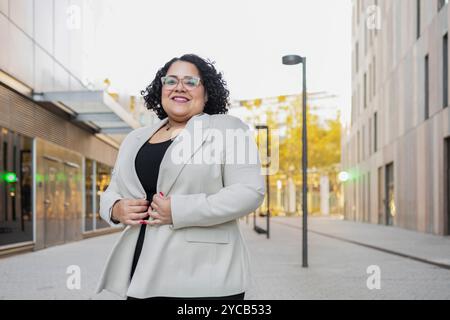 Une femme d'affaires hispanique aux cheveux bouclés, portant des lunettes et un blazer beige clair, sourit chaleureusement à la caméra. Elle se tient confiante dans un ur moderne Banque D'Images