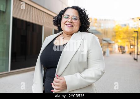 Une femme d'affaires hispanique confiante avec des cheveux bouclés, portant des lunettes et un blazer blanc élégant sur un haut noir, souriant et regardant l'appareil photo tout en Banque D'Images