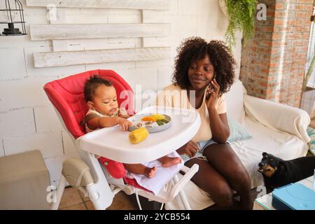 Une mère afro-américaine discute sur son mobile pendant que son bébé prend un repas dans une chaise haute, avec un petit chien qui regarde à proximité. La scène se déroule dans un c. Banque D'Images
