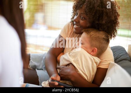 Une femme afro-américaine embrasse amoureusement un enfant caucasien assis sur un canapé, partageant un moment joyeux et tendre. La femme sourit chaleureusement au Banque D'Images