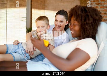 Une famille multiraciale joyeuse se détend sur un canapé, s'engageant joyeusement avec un smartphone. L'image montre une femme afro-américaine, une femme caucasienne, Banque D'Images
