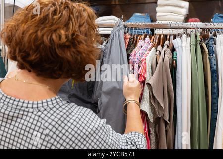 Une femme adulte trie à travers une variété de vêtements colorés suspendus sur un support de vente au détail dans un environnement de shopping, en se concentrant sur le processus de sélection Banque D'Images