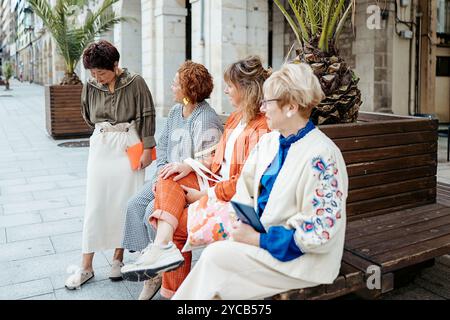 Quatre femmes de styles et d'apparences variés s'engagent dans une discussion animée tandis que trois d'entre elles sont assises sur un banc de rue, montrant une scène de frien Banque D'Images