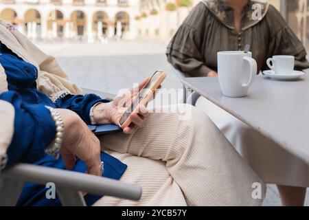 Cette image stock capture deux femmes matures engagées dans l'utilisation de leurs smartphones tout en étant assis à une table de café en plein air, ils sont habillés de façon décontractée et l'application Banque D'Images