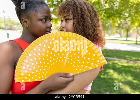 Deux jeunes femmes, une afro-américaine et une latino-américaine, partagent un moment tendre dans un parc, en utilisant un ventilateur aux couleurs vives pour plus d'intimité Banque D'Images