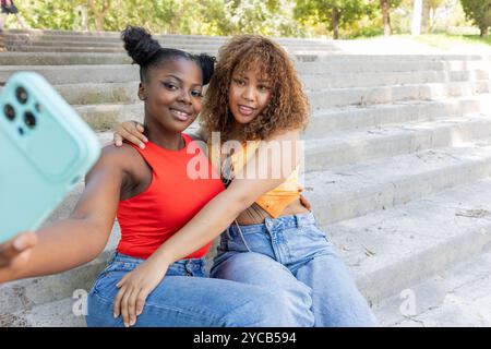 Les filles LGBTQIA+ afro-américaines et latino-américaines profitent d’une journée ensoleillée dans le parc et prennent un selfie ensemble sur les marches Banque D'Images