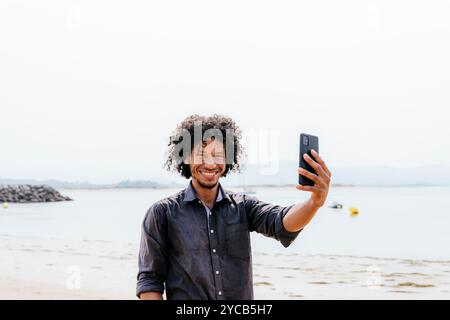 Un jeune homme joyeux aux cheveux bouclés prend un selfie sur son smartphone par le bord de mer son sourire et la toile de fond pittoresque des eaux calmes et du ciel clair en Banque D'Images