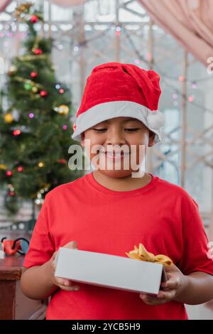 Garçon latino-américain souriant portant un chapeau de Père Noël et un t-shirt rouge, tenant une boîte cadeau avec un arc, avec un arbre de Noël et des lumières en arrière-plan Banque D'Images