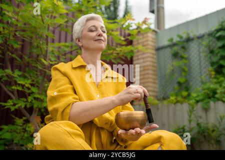 Une femme mature sereine aux cheveux blancs courts médite avec un bol chantant himalayen dans un jardin calme et luxuriant, les yeux fermés et vêtu d'un jaune vif Banque D'Images