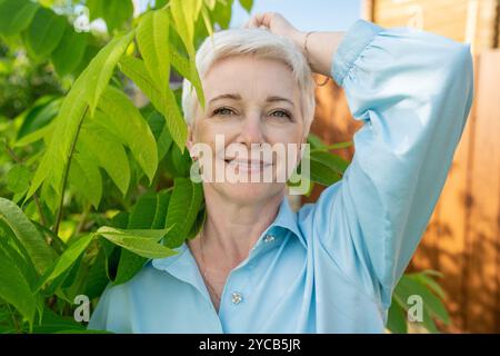 Une femme mature joyeuse aux cheveux blancs courts, portant une chemise bleu clair, sourit directement à la caméra, entourée de feuilles vertes vibrantes. Banque D'Images