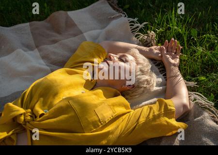 Sereine femme mature se trouve les yeux fermés sur une couverture dans un champ, le soleil illuminant son visage, exsudant une aura paisible dans un cadre naturel tranquille. Banque D'Images