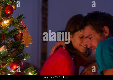 Un couple partage un moment intime près d'un sapin de Noël joliment décoré avec des lumières et des ornements lumineux, avec une décoration d'arbre en bois i. Banque D'Images