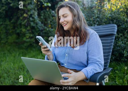 Gros plan portrait d'une femme jardinière souriante assise sur une chaise dans la serre, à l'aide d'un ordinateur portable et parlant sur le Web appel entouré de plantes exotiques. Domicile Banque D'Images