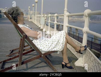 Années 1970, élégante dame mature en robe d'été et chaussures à talons, assise dans un transat en bois près des balustrades en fer sur une jetée côtière victorienne, Angleterre, Royaume-Uni. Banque D'Images