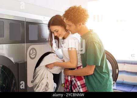 Jeune couple partageant un moment tout en faisant la lessive dans une laverie lumineuse Banque D'Images