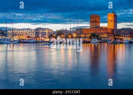 Vue imprenable en soirée sur l'hôtel de ville d'Oslo et Aker Brygge depuis les quais d'Akershus, avec des bâtiments illuminés se reflétant sur les eaux calmes sous un s spectaculaire Banque D'Images