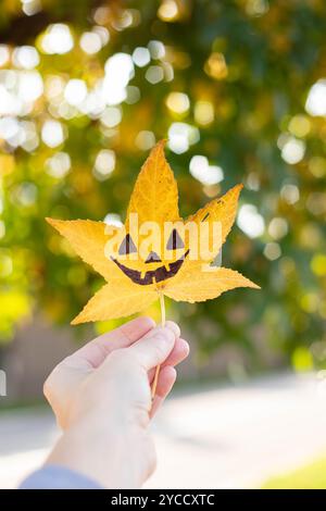 Main tenant une feuille de liquidambar jaune avec le dessin d'un visage de citrouille d'halloween dessus. Fond d'automne avec espace de copie. Prise de vue verticale. Banque D'Images