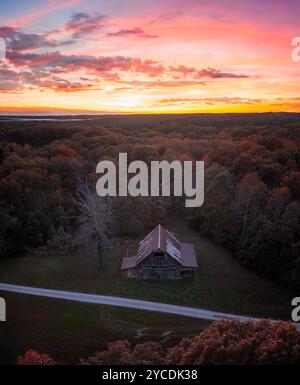 Une ancienne grange avec un toit en métal et un parement altéré se trouve dans une clairière dans les bois avec un coucher de soleil dynamique et coloré de l'Indiana au-dessus d'elle à l'horizon. Banque D'Images