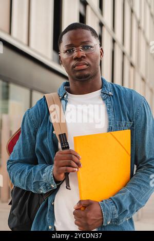 Vertical sérieux étudiant afro-américain regardant pensif à la caméra, tenant un dossier et portant un sac à dos, debout à l'extérieur dans une ville urbaine moderne, exprimant la confiance sur le campus universitaire. Photo de haute qualité Banque D'Images