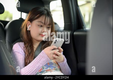 Une jeune fille asiatique adorable utilise son smartphone et écoute de la musique à travers ses écouteurs tout en voyageant avec ses parents, assis dans le dos Banque D'Images