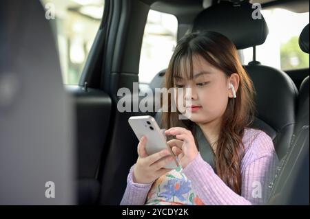 Une jeune fille asiatique adorable utilise son smartphone et écoute de la musique à travers ses écouteurs tout en voyageant avec ses parents, assis dans le dos Banque D'Images