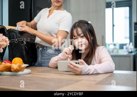 Une jeune fille asiatique regarde un dessin animé sur un smartphone tout en prenant le petit déjeuner avant l'école, avec sa mère emballant un sac à dos d'école à la salle à manger ta Banque D'Images