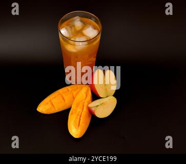 Un grand verre d'un mélange de jus de fruits avec de la glace sur un fond noir, à côté de morceaux de pomme mûre et de mangue. Gros plan. Banque D'Images