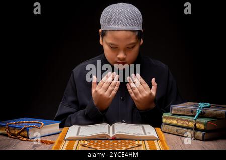 Un garçon musulman asiatique est assis et lit le Coran à la table en bois de sa maison pendant la nuit pendant le mois de ramadan, calligrap arabe Banque D'Images