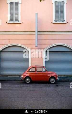 Cremona, Lombardie, Italie 15 octobre 2024 Une petite Fiat vintage rouge 500 est garée dans une rue calme devant un immeuble résidentiel rose pastel Banque D'Images