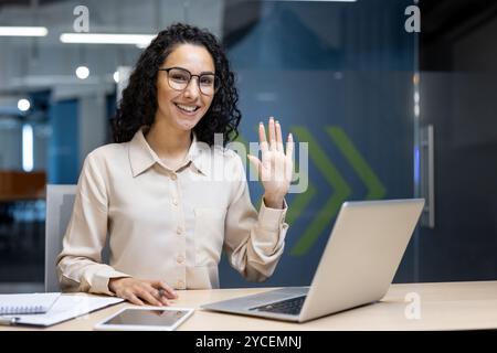 Femme hispanique au bureau agitant à la caméra, assise au bureau avec ordinateur portable, téléphone, documents. Reflète un environnement de travail amical, une interaction professionnelle, une communication positive, un comportement confiant. Banque D'Images