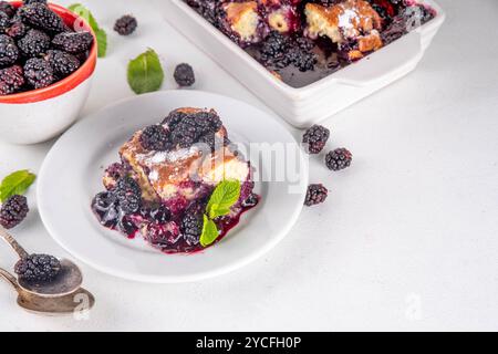 Tarte de cordonnier de mûrier maison sucrée, gâteau de cordonnier de baies d'été doux dans un plat de cuisson en émail et une assiette, sur un espace de copie de table de cuisine blanc Banque D'Images