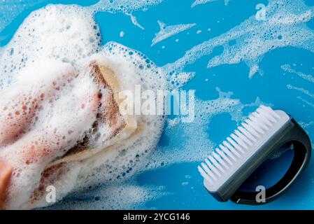 Main tient une éponge de nettoyage et essuie une mousse savonneuse sur un carreau bleu. fond bleu. Concept de nettoyage, service de nettoyage. Bannière. Plat, vue de dessus Banque D'Images