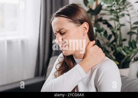 Triste jeune femme douloureuse avec douleur au cou mal de corps du travail, tenant massant frottant mal à l'épaule, mal assis sur le canapé à la maison. Douleur muscles du corps s Banque D'Images