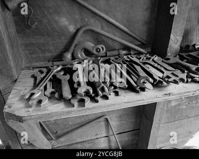 Grand groupe d'outils métalliques, principalement des clés ou des clés, empilés sur un établi en bois dans un hangar en bois, noir et blanc. Ambiance vintage. Banque D'Images