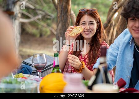 Heureuse jeune femme mangeant et partageant de la nourriture pendant le pique-nique en plein air. Moment de style de vie authentique naturel avec des amis profitant d'un rassemblement estival dans la nature Banque D'Images