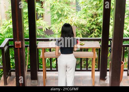 Une femme se tient debout à une table en bois dans un cadre extérieur verdoyant et luxuriant, entouré de plantes tropicales. Elle porte un haut noir et un pantalon blanc, avec elle Banque D'Images