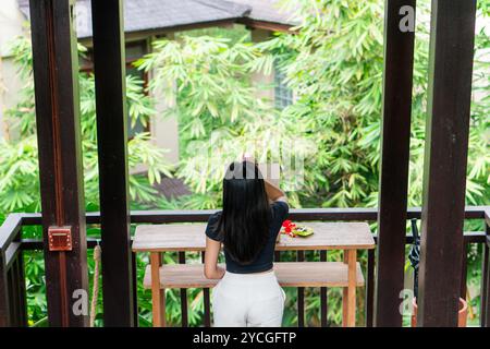 Une femme se tient debout à une table en bois dans un cadre extérieur verdoyant et luxuriant, entouré de plantes tropicales. Elle porte un haut noir et un pantalon blanc, avec elle Banque D'Images