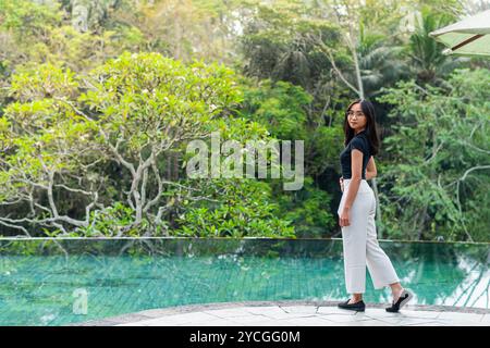 Une femme se tient près d'une piscine sereine entourée d'une végétation luxuriante. Elle porte un haut noir et un pantalon blanc, regardant l'eau tranquille Banque D'Images