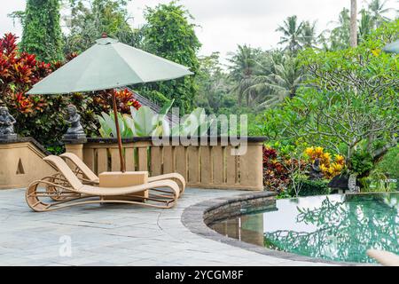 Une piscine extérieure sereine avec une chaise longue en bois avec un parasol vert. Des plantes tropicales luxuriantes et des arbres entourent la piscine, créant un tranqui Banque D'Images
