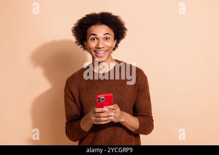 Joyeux jeune homme en pull brun tenant le téléphone rouge sur fond beige Banque D'Images