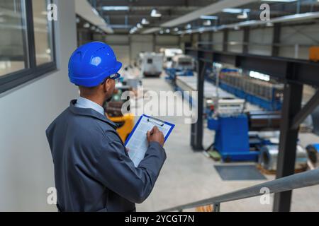 Homme afro-américain avec un casque bleu dans une unité d'usine, un ingénieur regardant des graphiques sur une tablette vérifiant un processus d'automatisation, vue de côté. Banque D'Images