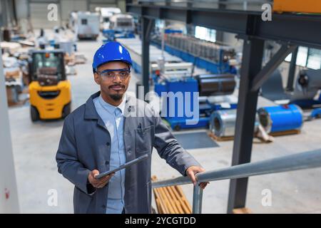 Homme afro-américain avec un casque bleu dans une unité d'usine, un ingénieur regardant des graphiques sur une tablette vérifiant un processus d'automatisation, vue de côté. Banque D'Images