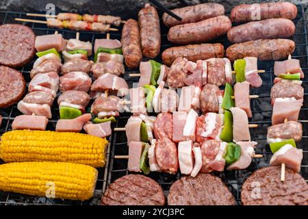 Gros plan de diverses viandes et légumes sur le gril barbecue. Saucisses crues, galettes de bœuf, kebabs et maïs grillés pendant la cuisson en extérieur préparati Banque D'Images