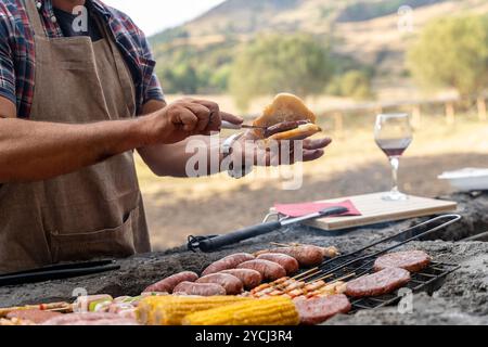 Chef préparant des hamburgers et des saucisses sur barbecue en plein air dans un cadre de montagne. Préparation naturelle des aliments pendant le barbecue d'été avec des ingrédients frais Banque D'Images